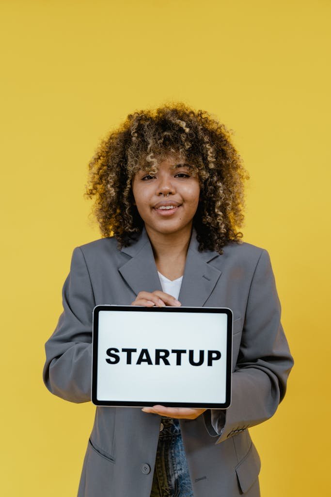 Smiling woman with curly hair holding a tablet displaying 'STARTUP' against a yellow background.